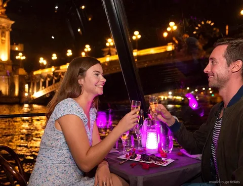 Couple toasting with champagne on a night Seine dinner cruise, a lit bridge behind, before the Moulin Rouge tickets show
